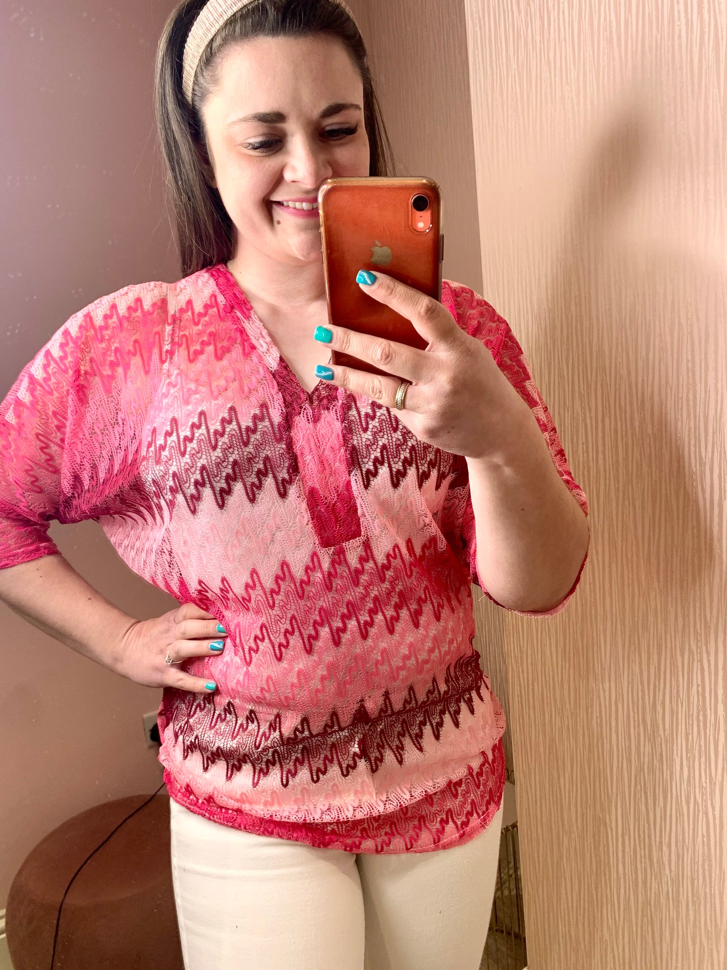Young brunette female stood in front of a pink wall and taking a photo of herself on her phone in the mirror.  She is wearing white skinny trousers and a crochet knit v-neck top.  It has three quarter length sleeves and is a zig zag knit with differing shades of pink.  Her free hand is on her hip.  She is wearing a pink headband and smiling.