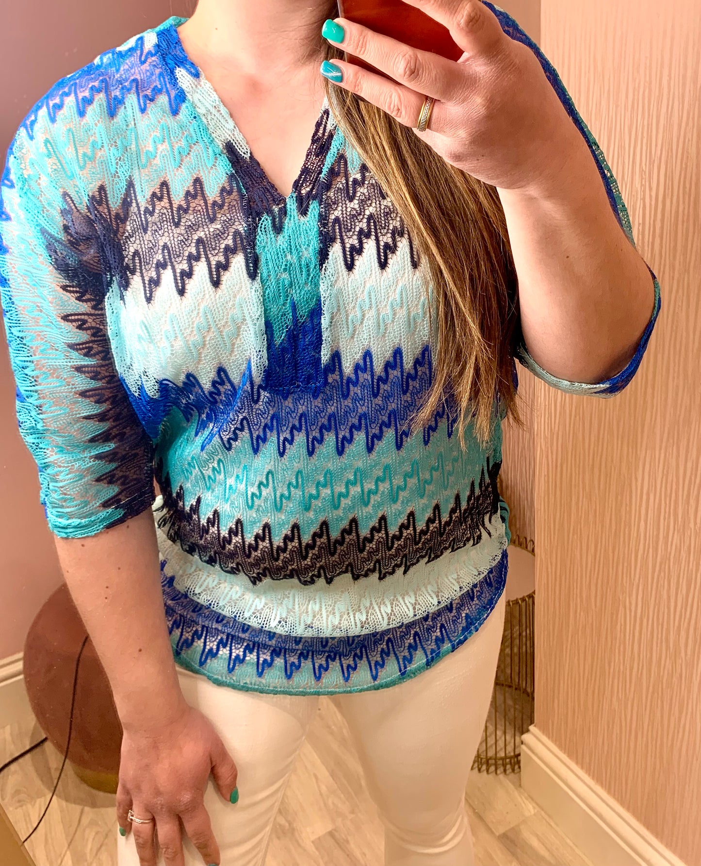Close up of a female stood in front of a pink wall and taking a photo of herself on her phone in the mirror.  She is wearing white skinny trousers and a crochet knit v-neck top.  It has three quarter length sleeves and is a zig zag knit with differing shades of blue.  Her free hand is on her thigh.