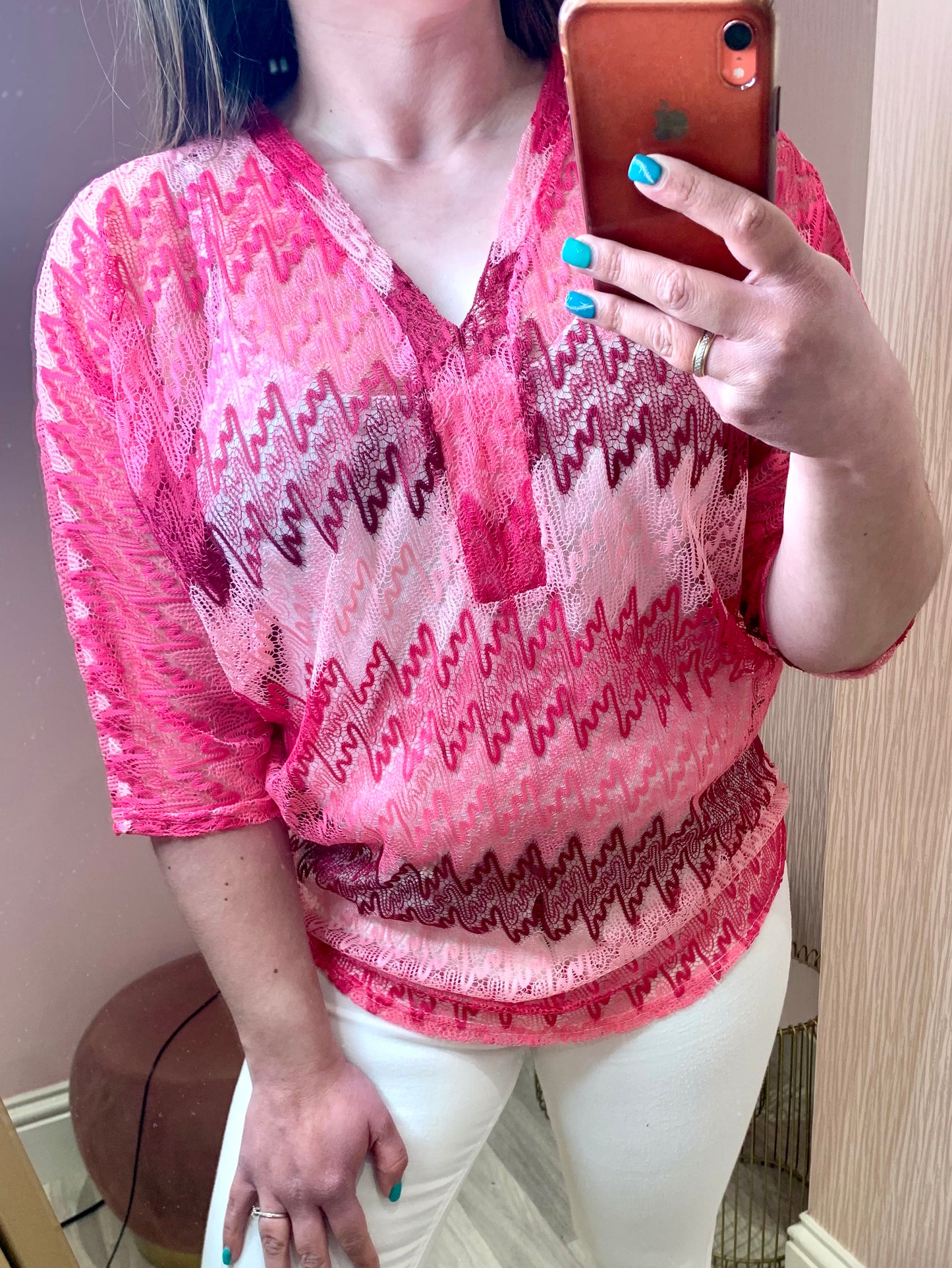 Close up of a female stood in front of a pink wall and taking a photo of herself on her phone in the mirror.  She is wearing white skinny trousers and a crochet knit v-neck top.  It has three quarter length sleeves and is a zig zag knit with differing shades of pink.  Her free hand is on her thigh.