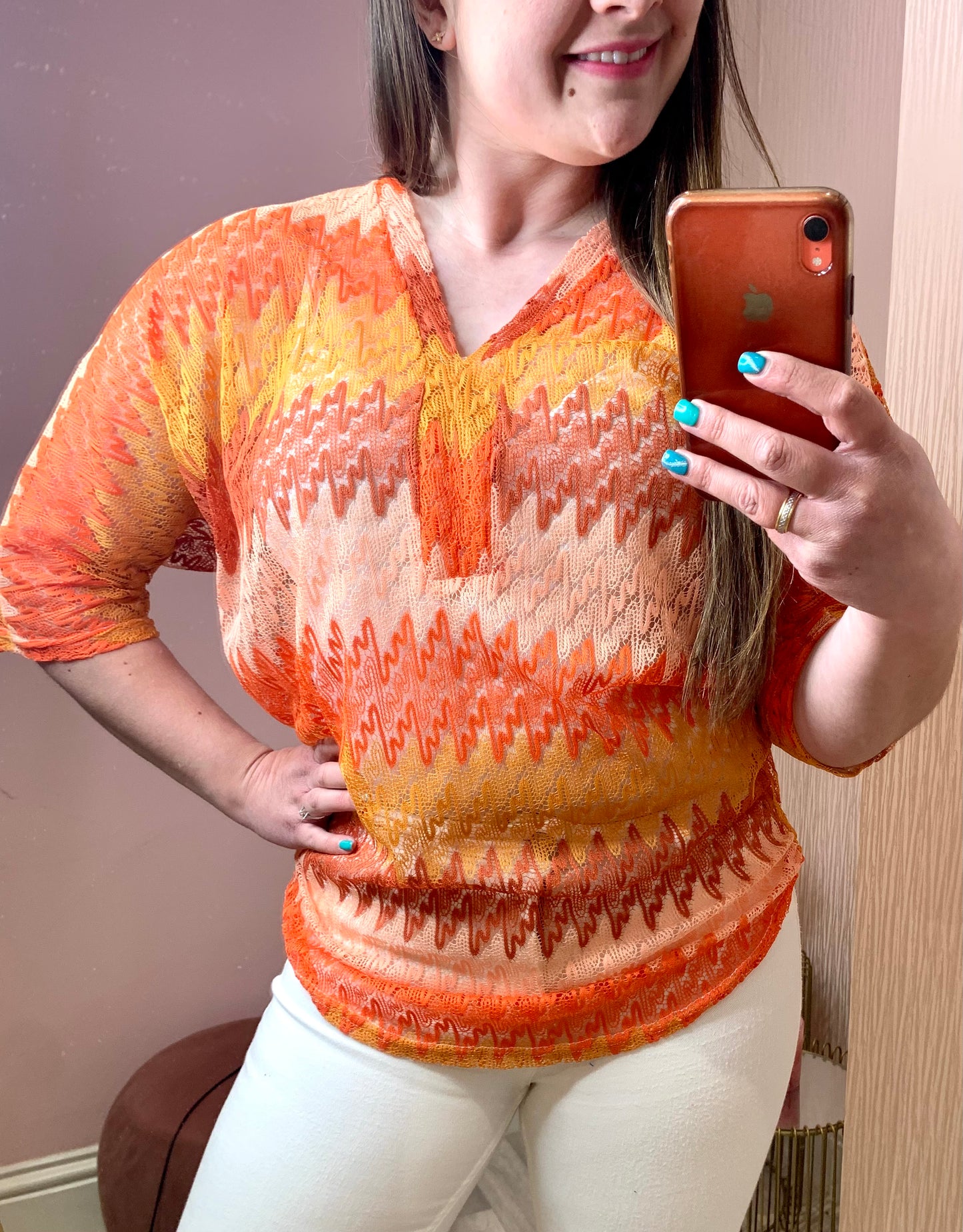 Close up of a young bruntette female stood in front of a pink wall and taking a photo of herself on her phone in the mirror.  She is wearing white skinny trousers and a crochet knit v-neck top.  It has three quarter length sleeves and is a zig zag knit with differing shades of orange.  Her free hand is on her hip.